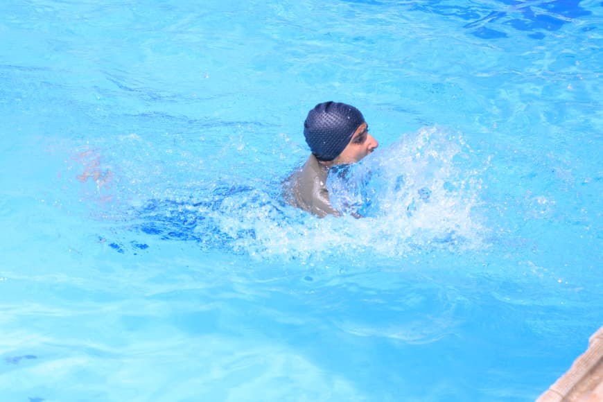 Swimmer wearing a black swim cap treading water in a bright blue pool with splashes nearby.