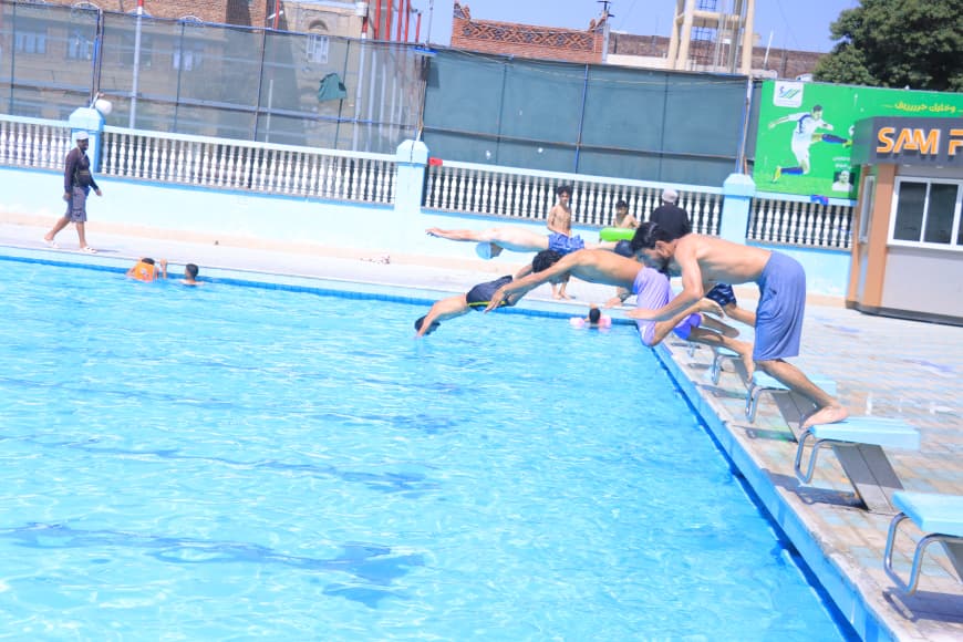 Two male swimmers dive off starting blocks into a bright blue pool as onlookers watch nearby at a public pool facility.