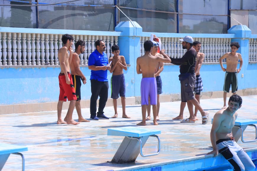 Coach giving instructions to a group of boys by a poolside near a blue wall.