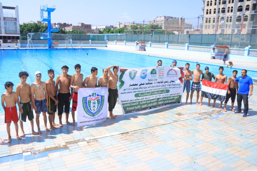 Group of boys in swimwear lined up along the edge of a large outdoor pool, holding a banner with Arabic text and logos.