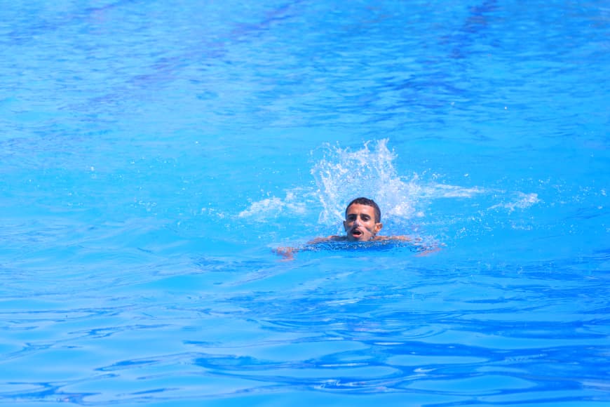 Man swimming in bright blue water, head above the surface with a splash around him, as he moves forward.