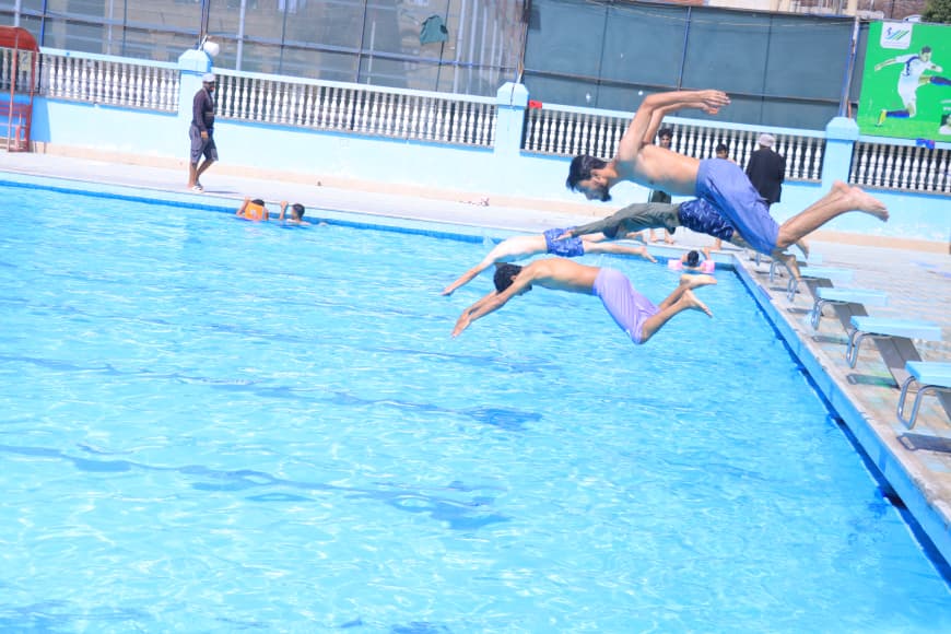 Three male swimmers are mid-dive into a bright blue pool from the edge, water splashing as they jump.
