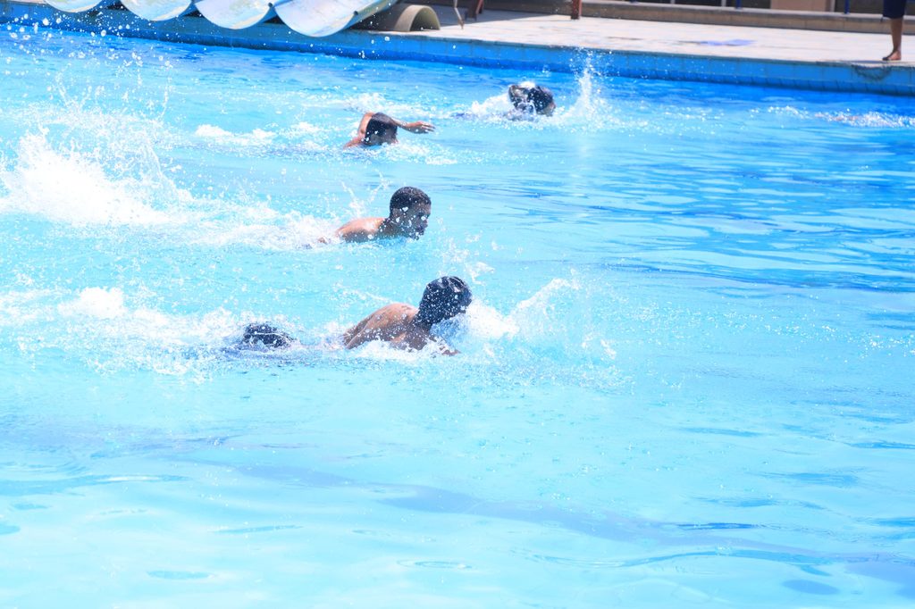 Multiple people swim across a bright blue outdoor pool, splashing water as they move.
