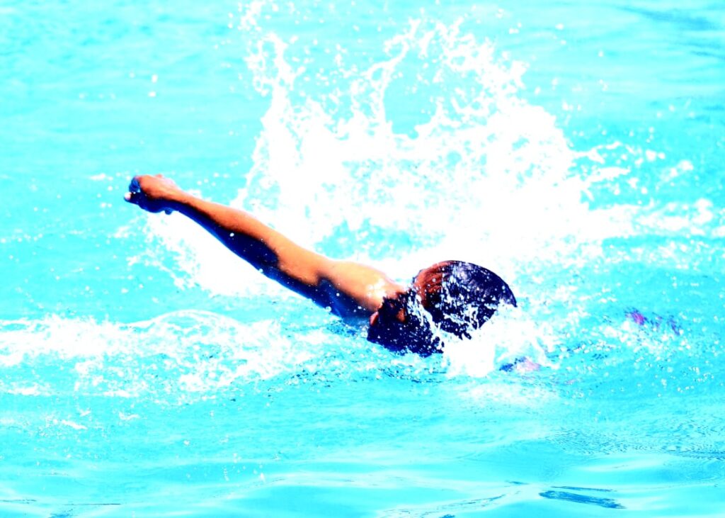 Swimmer mid-stroke in a bright blue pool, arm extended forward as water splashes around.