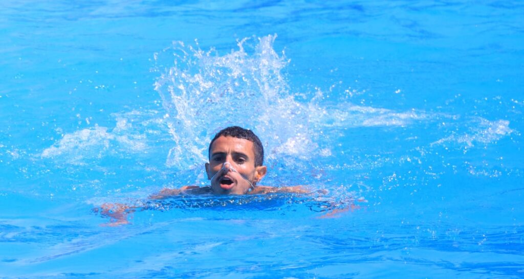 Man swimming in a bright blue pool with a snorkel mouthpiece, water splashing around him in front of the camera.