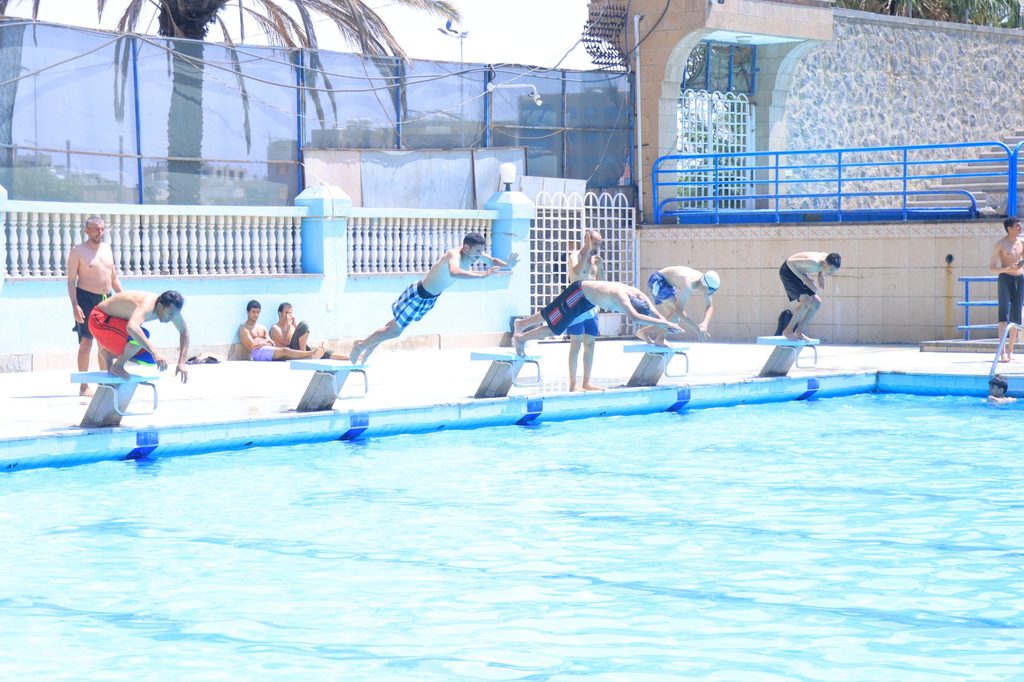 People on diving blocks at an outdoor pool, ready to dive into bright blue water as others watch nearby.