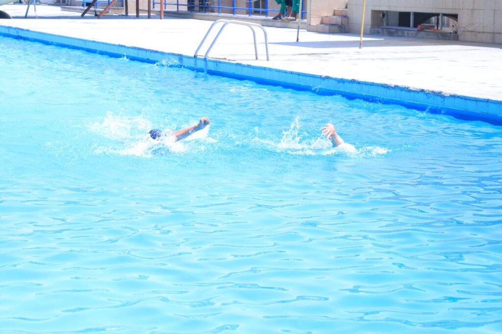 Two swimmers splash in a bright blue pool by the edge of a concrete deck with a metal ladder nearby.