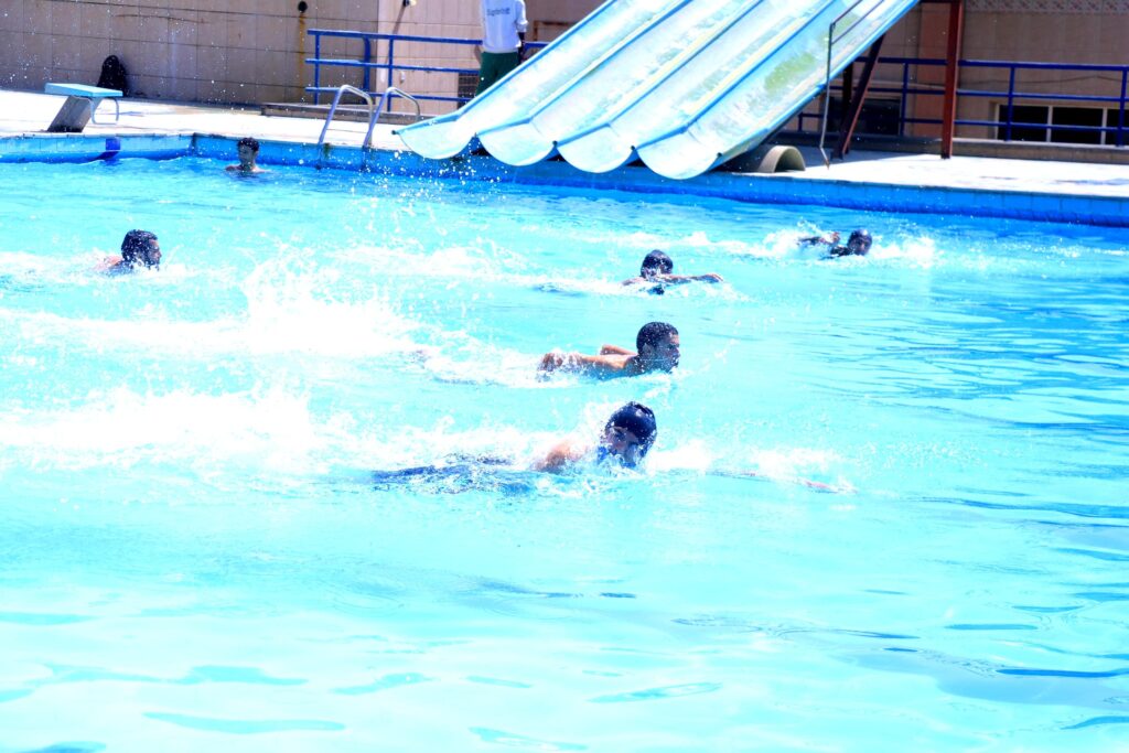 Children swimming and splashing in a bright blue pool with a large blue slide in the background.