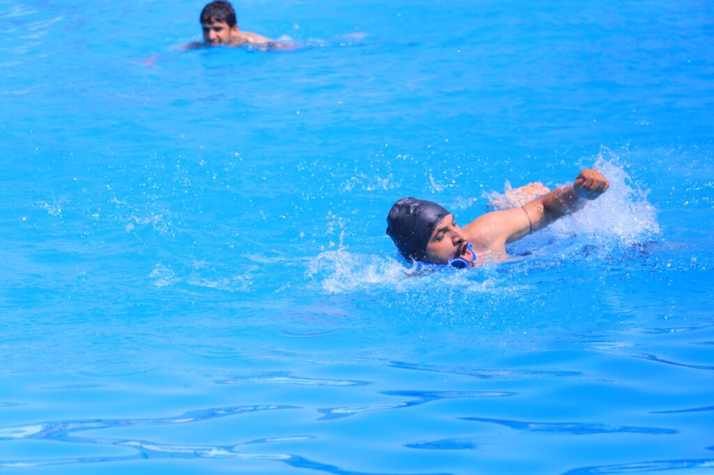 Swimmer wearing a black cap and blue goggles in a bright blue pool, arm extended as he swims; another swimmer visible in the background.