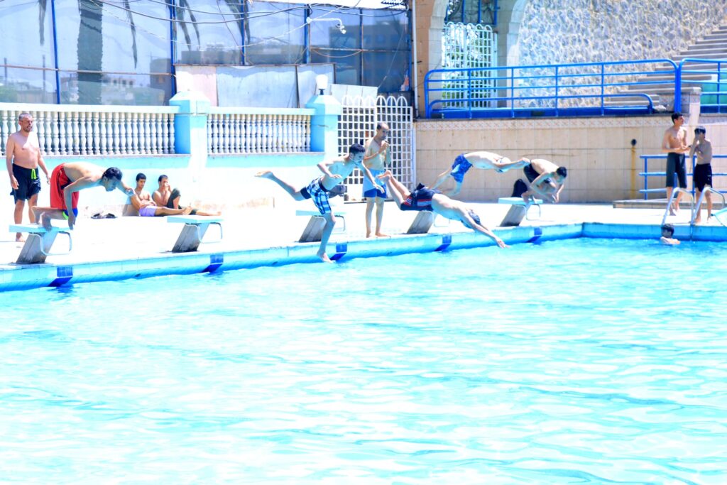 Several boys dive into a bright blue outdoor pool from starting blocks as onlookers stand poolside.