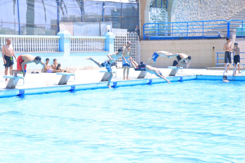 Group of boys diving off starting blocks into a sunlit outdoor pool while others sit and watch nearby.
