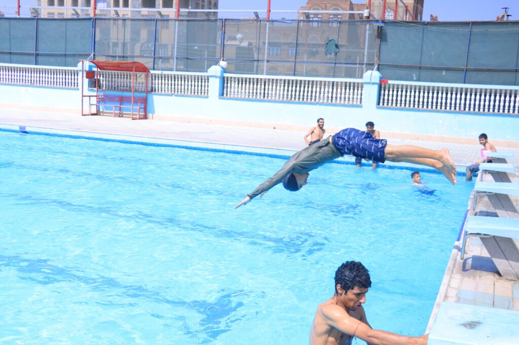 Person performs a dive into a bright blue outdoor pool while others stand at the edge watching.