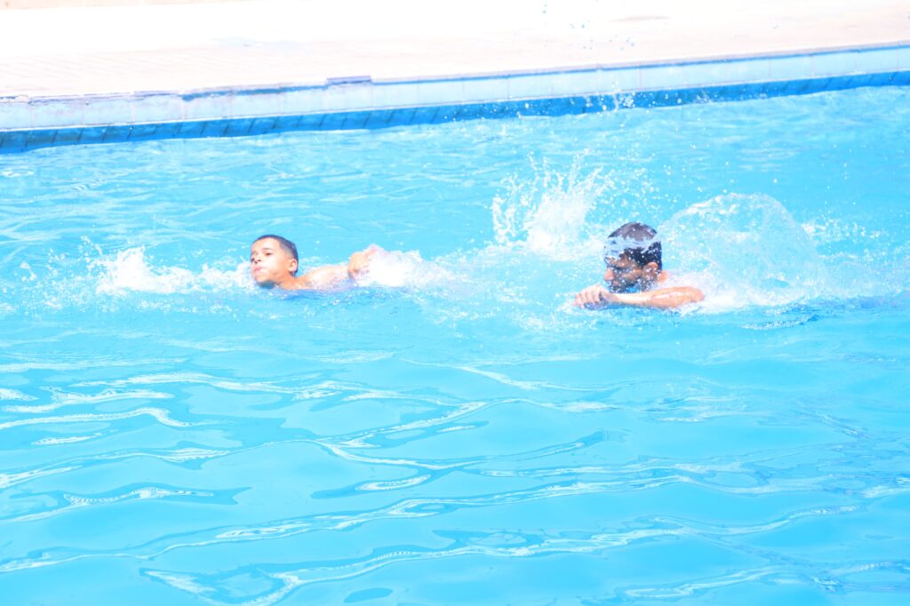 Two people swimming in a bright blue pool with splashes near the edge of the pool deck.