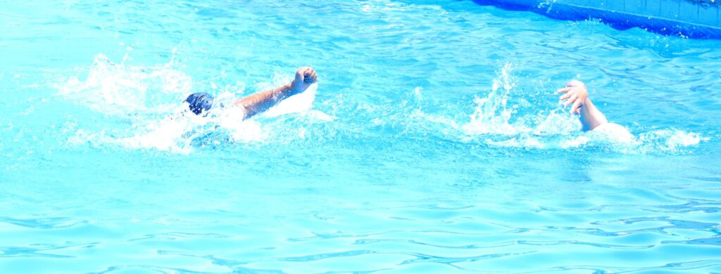 Two swimmers in a bright blue pool, mid-stroke with splashes around as one arm reaches forward and the other arm trails behind.