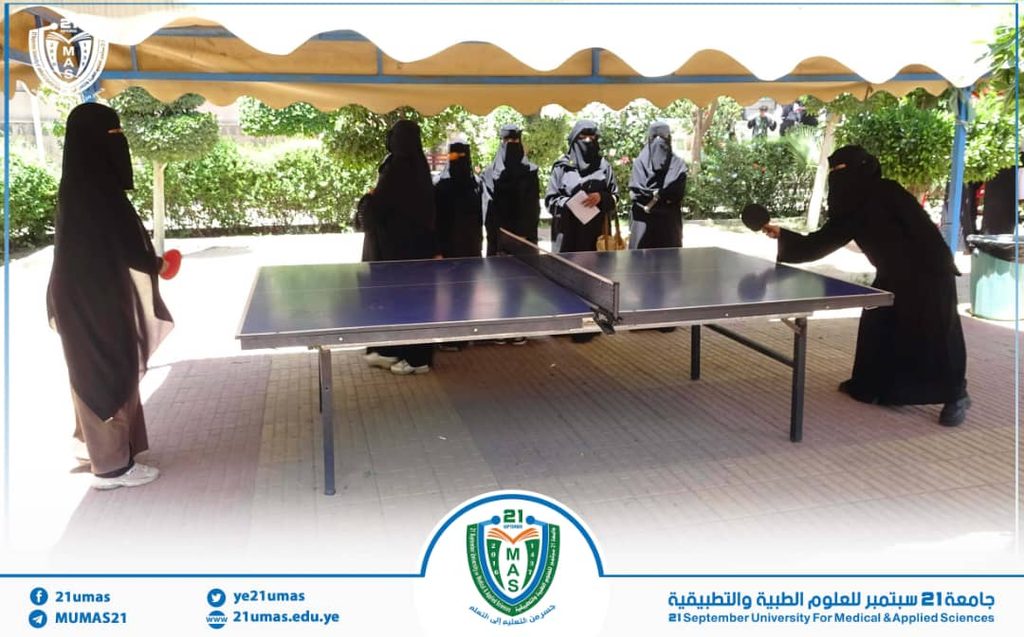Group of women in black abayas watch a table tennis match under a canopy in an outdoor university area.