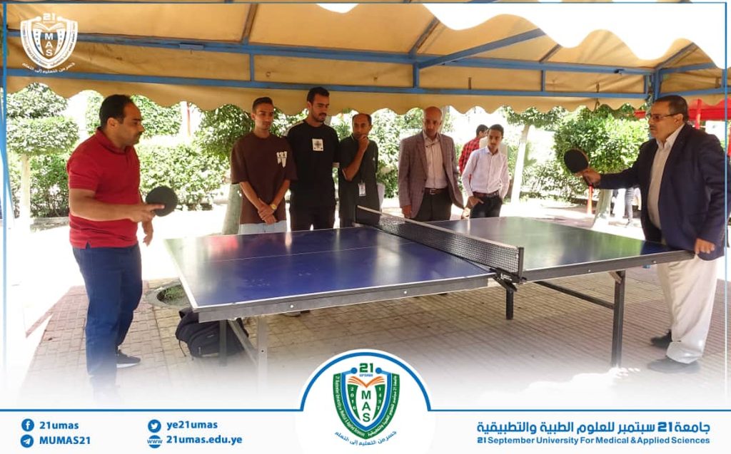 Group of men watch a man in a red shirt play table tennis under a sun canopy at an outdoor university event or gathering.