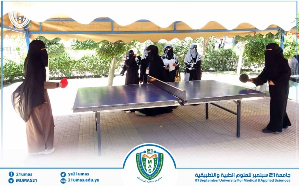 Group of women in abayas playing table tennis outdoors under a canopy at 21 September University.