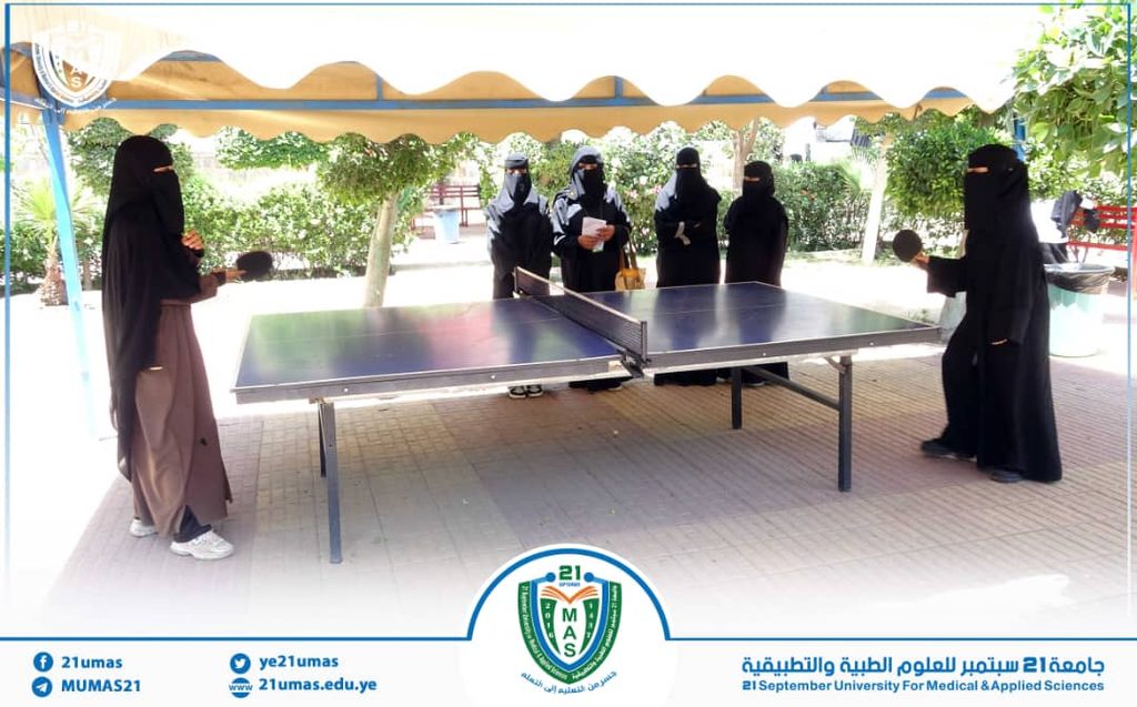 Women in abayas and niqabs playing table tennis outdoors under a canopy at a university campus, with banners and a logo visible in the foreground.