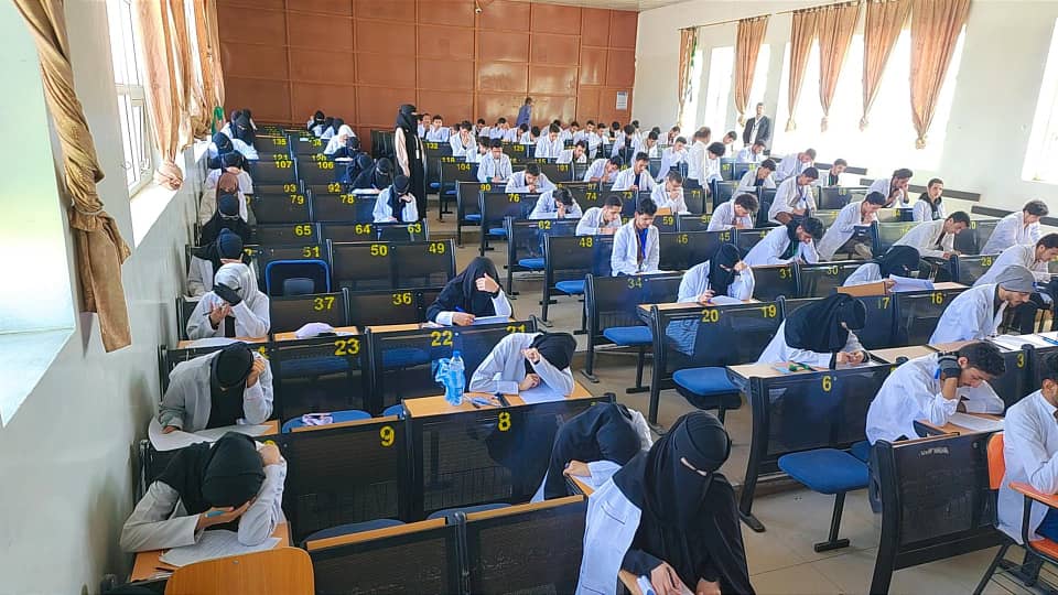 Crowded exam hall with students at numbered desks, heads down as they write.