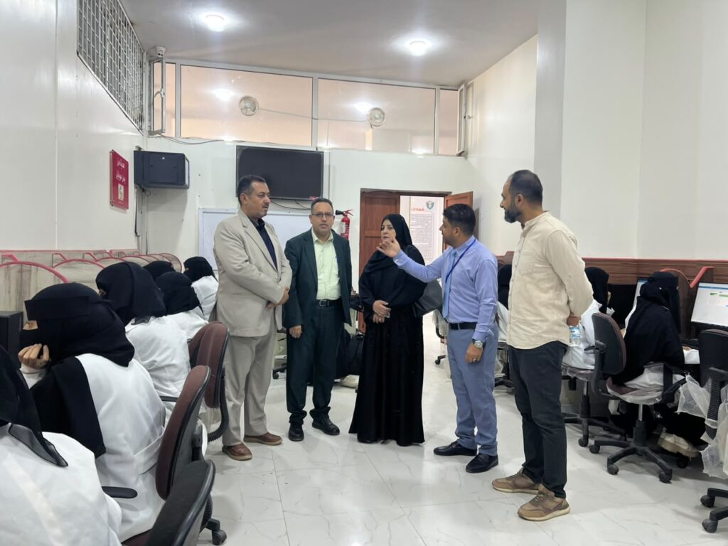 Five adults conversing in a computer lab while students in white coats sit at desks.