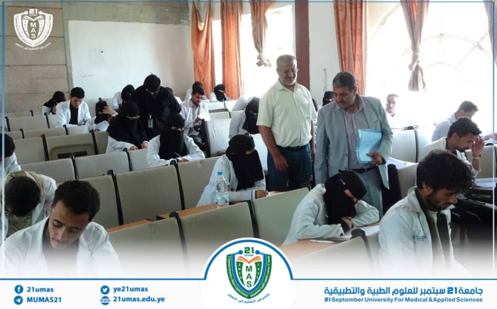 Exam hall with students in white coats taking a test; two men stand at the front, one holding a blue folder, logo banner at bottom.