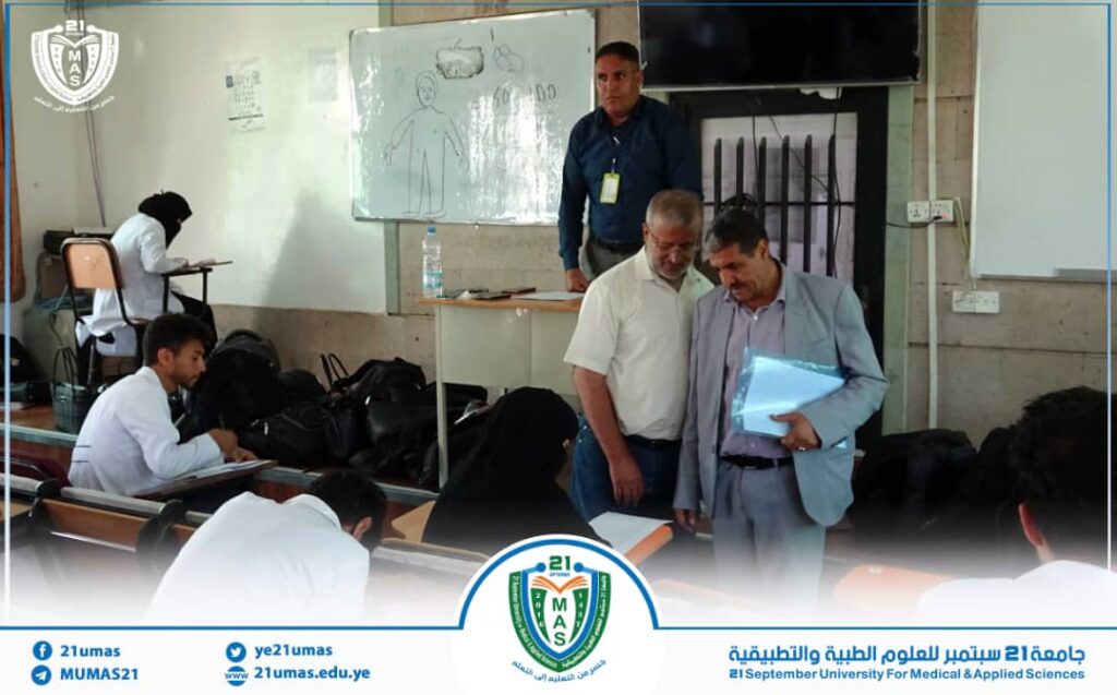 Classroom at 21 September University for Medical & Applied Sciences: two men review a folder while students study nearby.