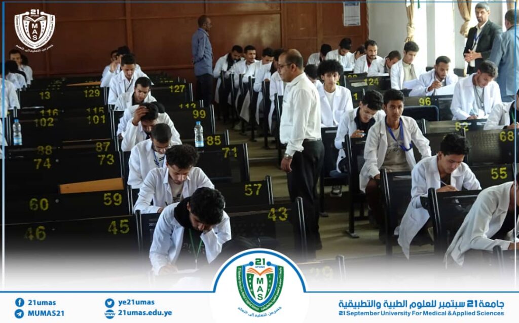 Medical students in white coats seated in rows, taking an exam in a large lecture hall with supervisors walking among them.