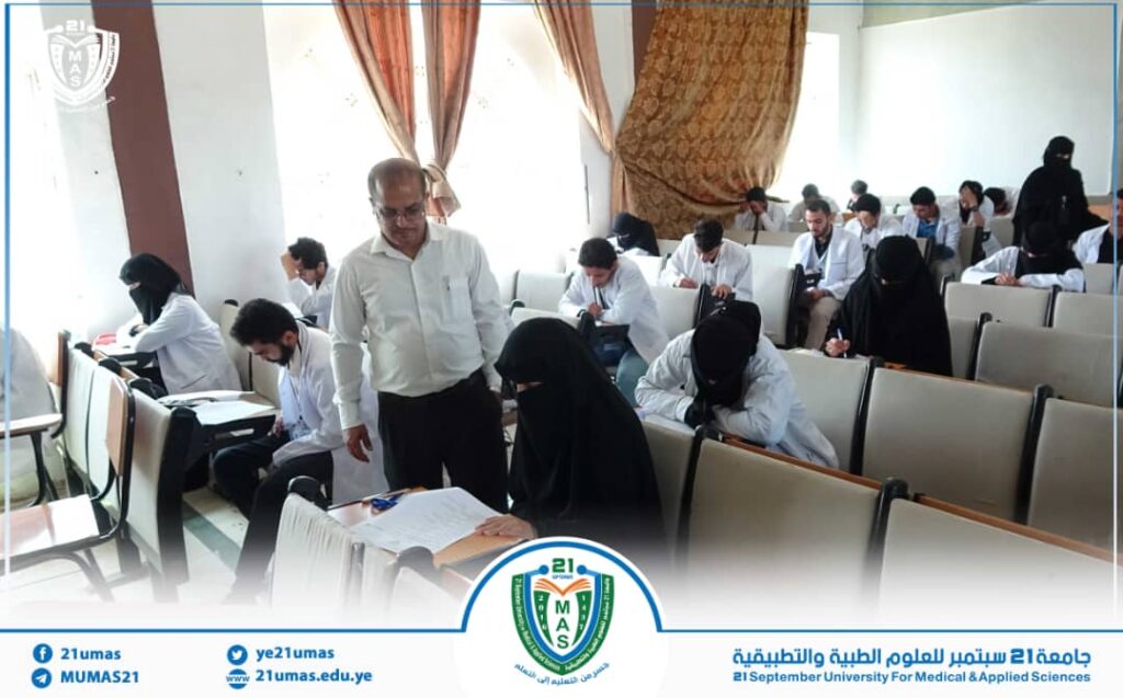 Professor overseing a university exam hall as students in white shirts and hijabs write at desks, some standing and reviewing papers ahead of the session.