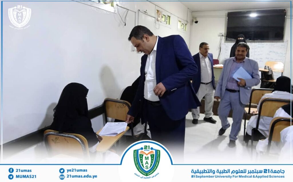 A man in a blue suit helps a seated woman in a hijab with paperwork in a classroom during a university event, with other men standing nearby and a university crest on the image frame.