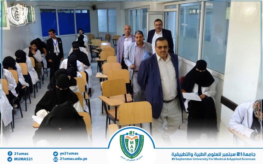 Group of men and women walking through a university classroom with empty desks, banner and logo visible at the bottom."Container scene shows attendees entering a lecture hall with a blue-accent banner and institutional emblem."