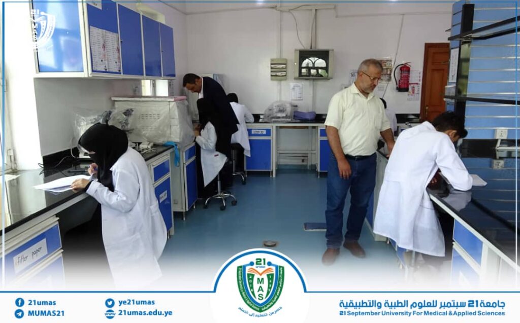 A university science lab with students in lab coats working at benches; an instructor stands nearby observing, with blue cabinets and equipment along the walls and a campus logo at the bottom center.