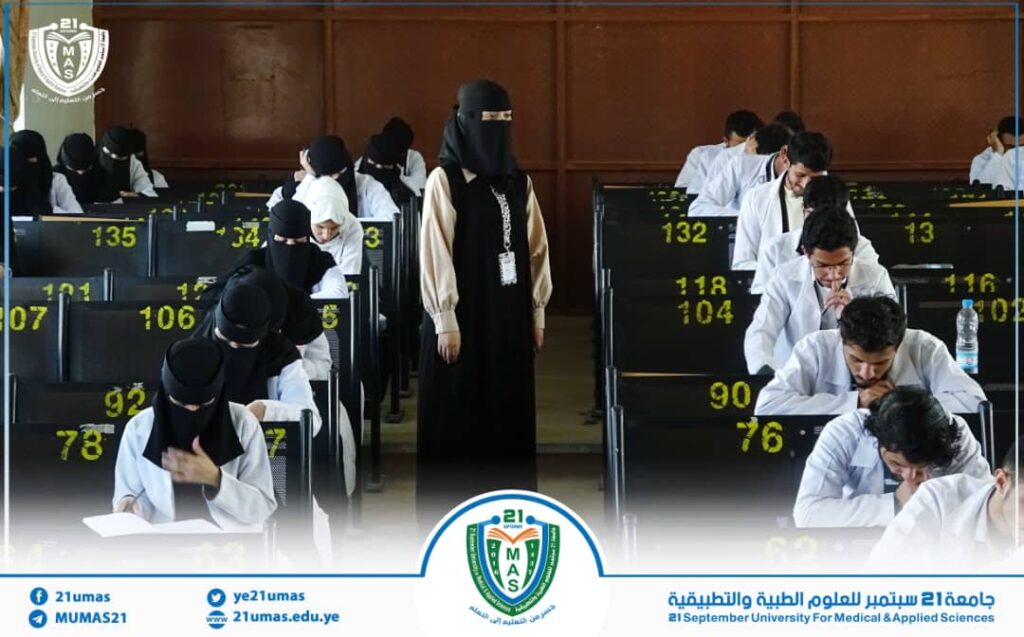 Exam hall scene: students in white coats taking written tests while a supervising woman in a black abaya oversees the room, with bright yellow seat numbers visible on chairs and a university banner at the bottom notch of the image.