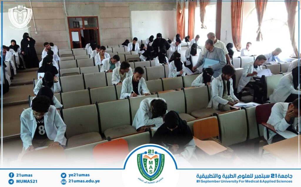 Students in white coats sit in rows in a lecture hall, taking an exam as a staff member distributes papers; university banner and logo visible at the bottom.