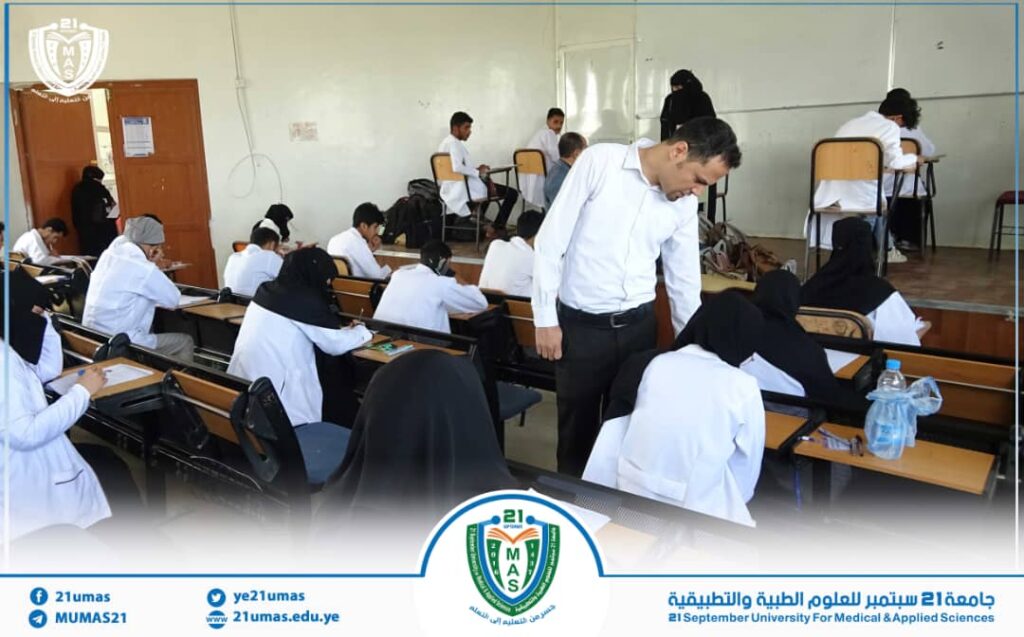 Students in white coats take an exam in a classroom as a male instructor leans over to help a student at their desk.