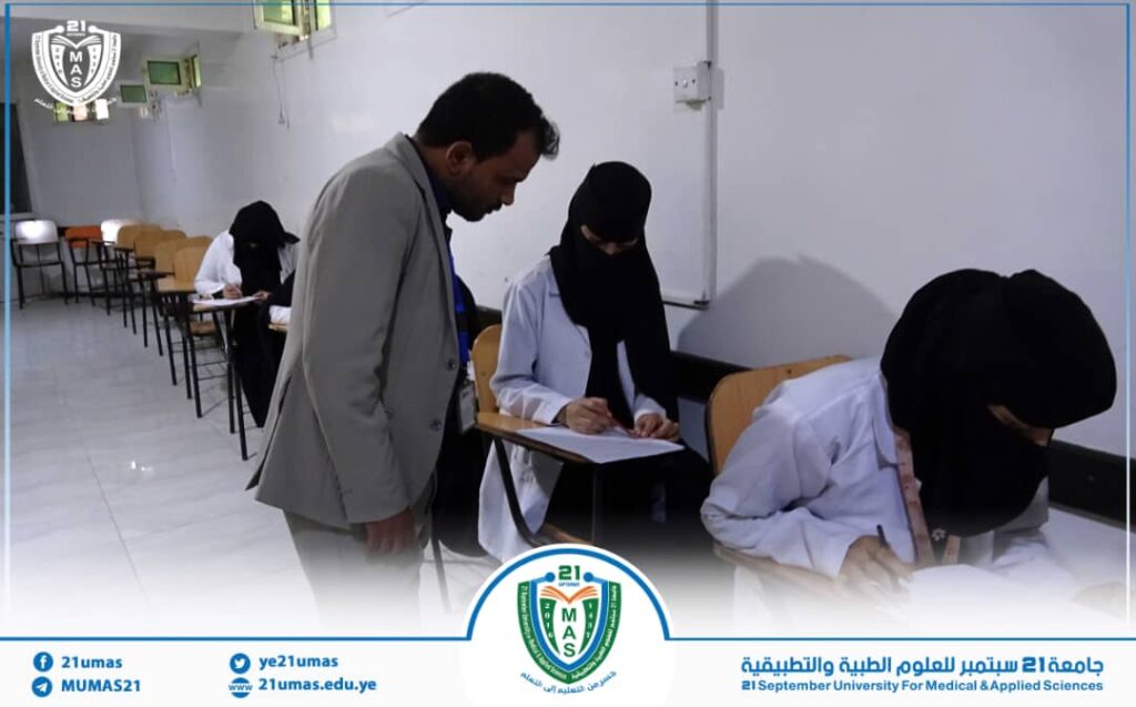 Instructor assists a hijab-wearing student taking notes at a desk during an exam in a classroom, with other students in the background.