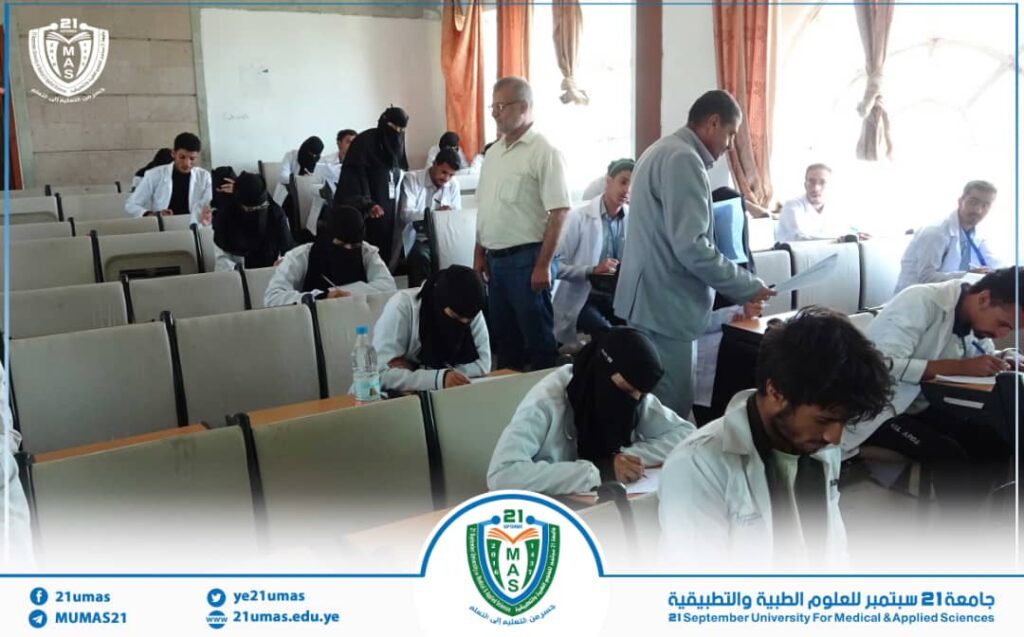 Medical students in white coats taking notes in a classroom; a man stands near the center as an instructor/supervisor, with a university logo and banner displayed at the bottom of the image.