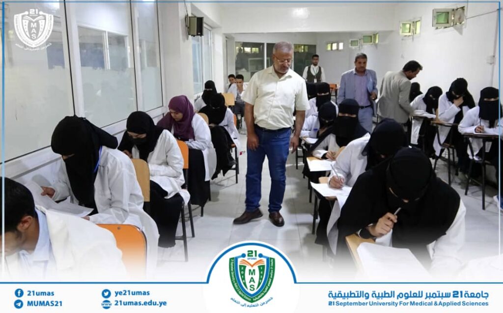 Exam hall at the 21 September University for Medical and Applied Sciences, with students in white coats and hijabs taking a written test while a supervisor monitors from the center.