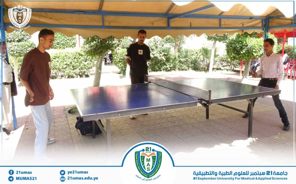 People play table tennis under a yellow tent at a university event, with logos on the tent and banner at the bottom.
