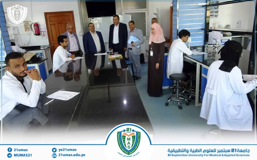 Group of eight people, including men in suits and a woman in a hijab, standing around a glossy lab counter while two lab technicians in white coats work at stations in a medical teaching lab at a university facility.