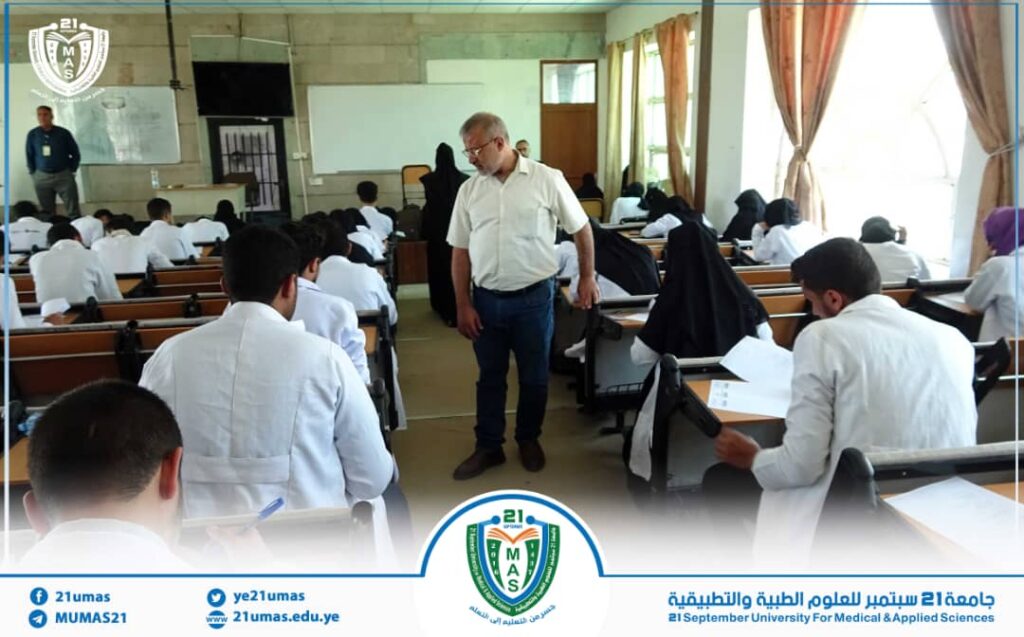 Instructor walks among students in a classroom filled with desks and white-coated attendees, university banner and crest visible at the front.