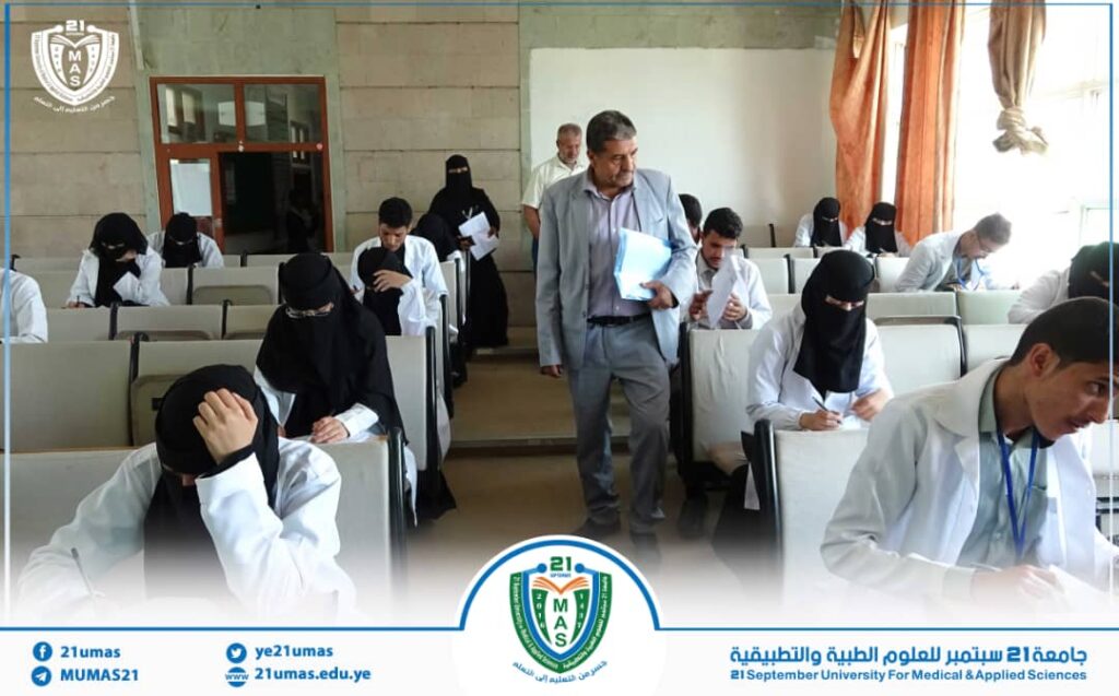 University classroom: students in white coats sit at desks, many wearing black hijabs, as a man in a gray suit walks between rows holding papers. A university banner and logo are visible at the front.