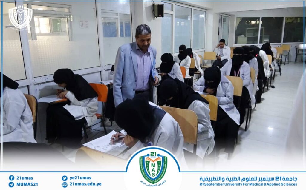 Lecturer walks between rows of female students in white coats taking an exam in a university classroom.