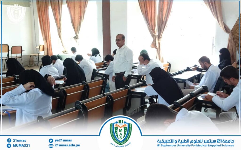 Male instructor stands at the front of a classroom as students in white shirts take notes around the desks.