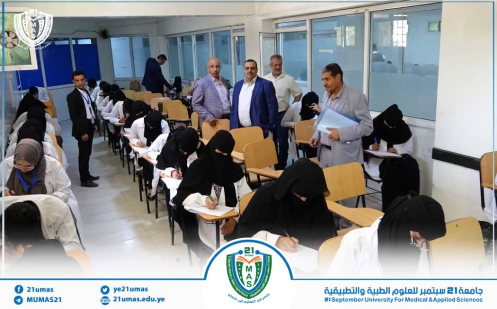 Students in hijabs seated in rows taking a written exam in a bright classroom, with several men standing at the back and a university logo at the front.