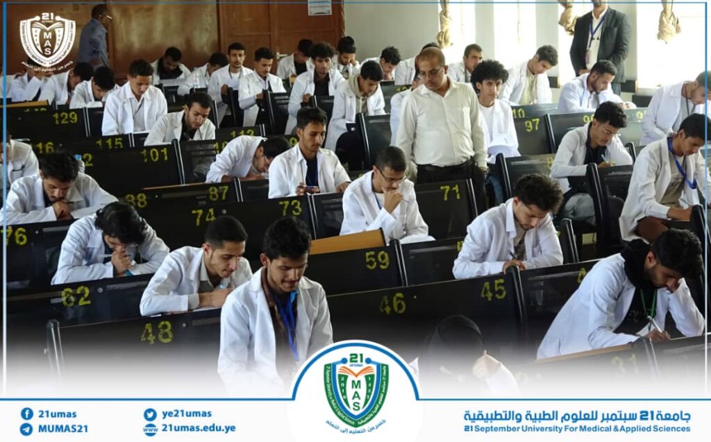 Lecture hall filled with students in white coats taking notes, a university crest in the upper left and a blue banner at the bottom.