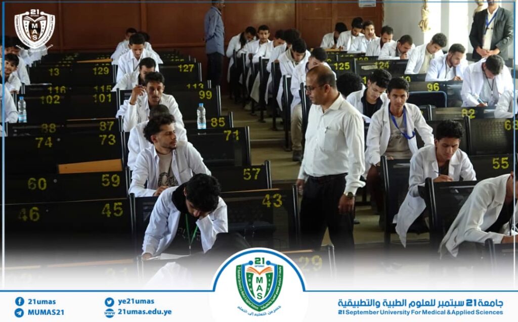 Group of students in white coats sit at numbered benches in a lecture/exam hall, with a supervising staff member walking through. University banner and logo visible at the bottom.