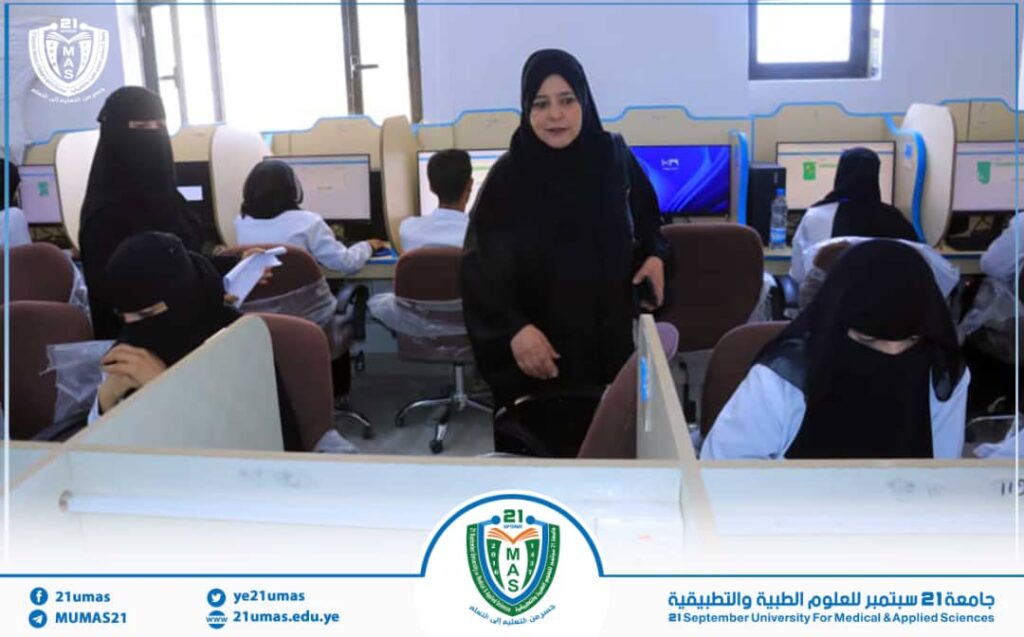 In a university computer lab, students sit at desks using desktop computers while a woman in a black abaya stands in the center of the room. A university crest is visible in the scene.
