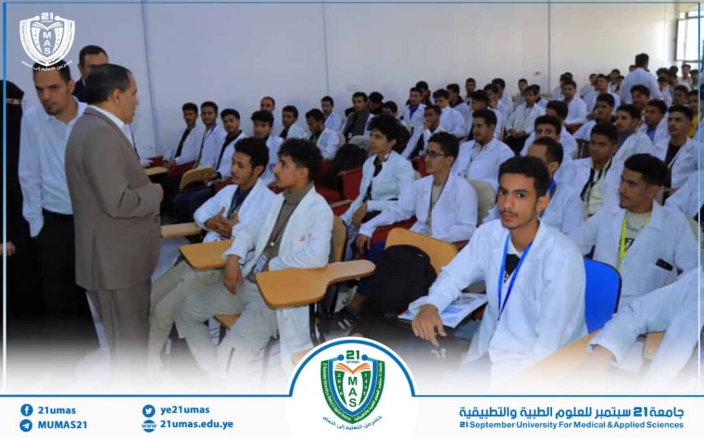 Lecture hall with medical students in white coats listening to a speaker at the front, university logo visible in the corner.