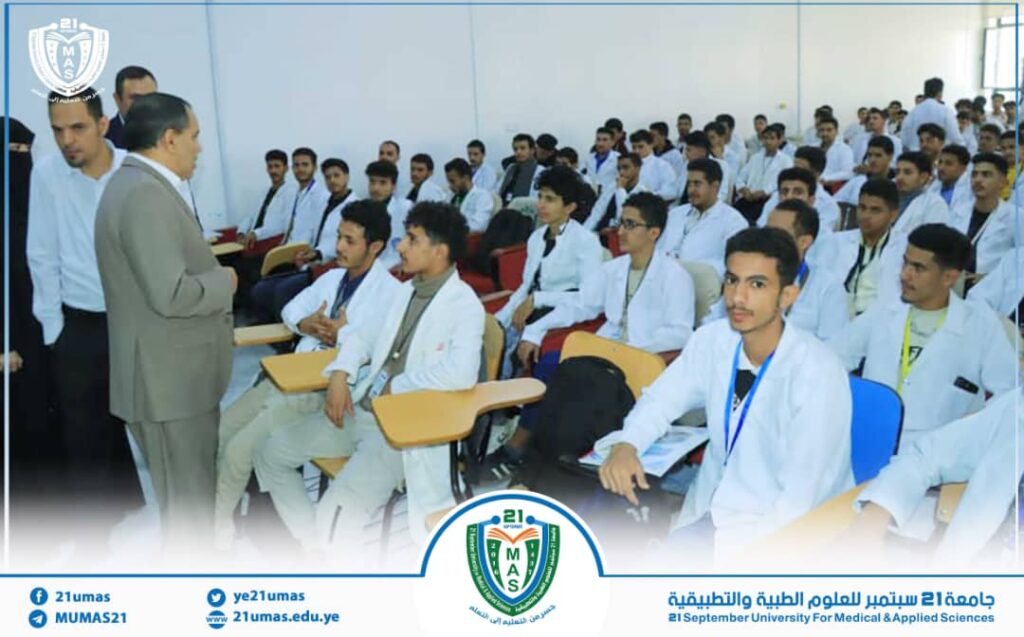 Instructor speaks to a large group of medical students in a classroom; students wear white coats and sit at desks as the lecturer stands at the front.