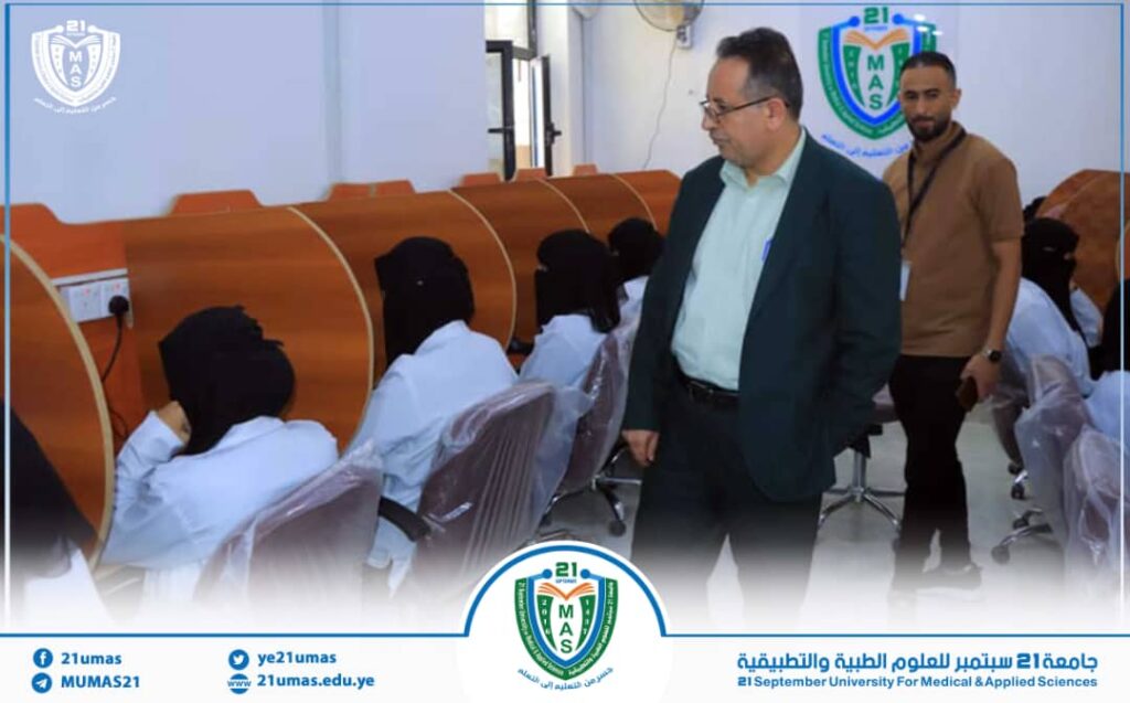 University official in a suit walking past students seated at study cubicles in a classroom campus setting with university logos visible